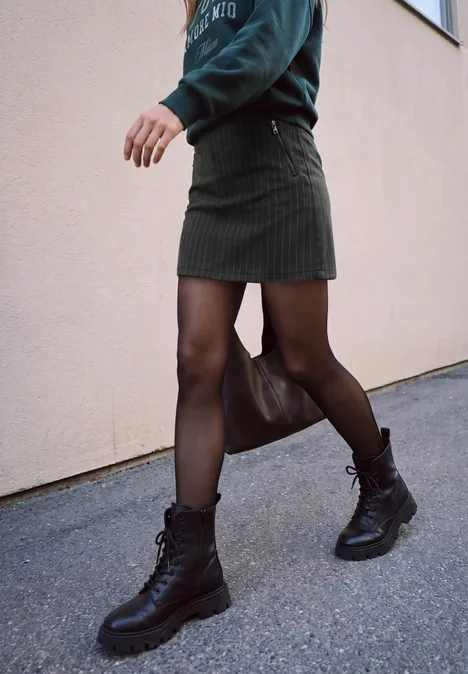 Minirock mit Elastikbund und Gallonstreifen Black Eine Person in schwarzer Strumpfhose, kurzem Rock und Boots läuft an einer Hauswand entlang.