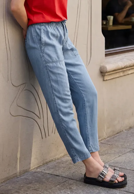 Person lehnt an Wand, trägt hellblaue Bundfalten-Hose, rotes Top und schwarze Sandalen mit Schnallen.