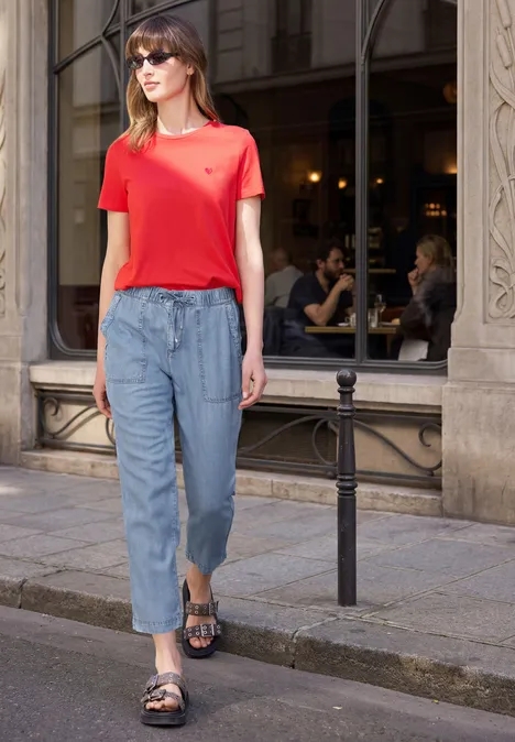Frau in rotem T-Shirt und blauen Jeans schlendert auf dem Bürgersteig vor einer Straßencafé-Fassade.