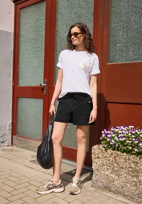 High Waist Shorts mit Knopfdetail Black Frau in weißem T-Shirt und schwarzen Shorts steht vor einer Haustür und hält eine schwarze Tasche.