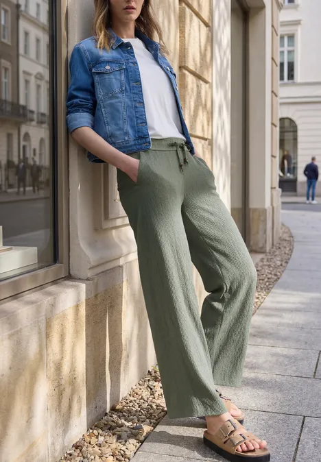 Eine Frau lehnt entspannt an einer Fassade, trägt Jeansjacke, weiße Bluse, grüne weite Hose und Sandalen.