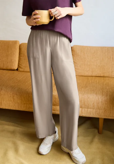 Wide Leg Culotte im Loose Fit quiet ivory Person trägt bequeme beige-weite Hose, lila Oberteil und weiße Sneaker, hält eine Tasse vor Sofa.