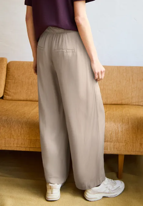 Wide Leg Culotte im Loose Fit quiet ivory Frau von hinten in beigen, weit geschnittenen Hosen und Sneakern, steht vor einem ockerfarbenen Sofa.