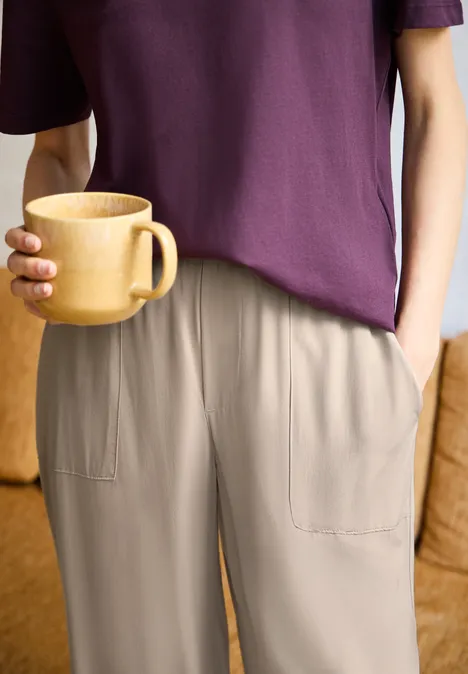 Wide Leg Culotte im Loose Fit quiet ivory Person in lila T-Shirt und beigen Hosen hält einen großen Holzbecher, eine Hand in der Tasche.
