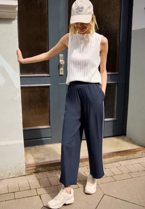 Frau in weißem ärmellosem Top, navy Culottes, weißen Sneakern und Baseballcap lehnt an Tür.