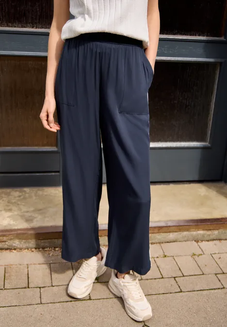 Person trägt weite dunkelblaue Hose, weiße Sneaker und helles Top, stehend vor Tür.