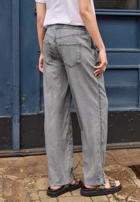 Eine Person steht vor einer Tür und trägt graue gerade geschnittene Jeans mit Sandalen.