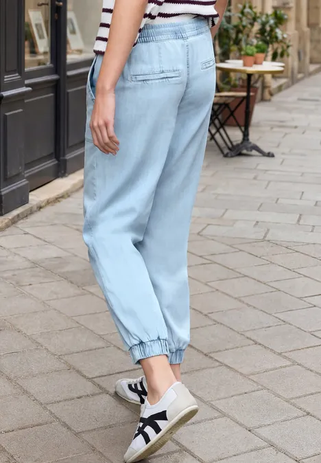 Persoon draagt losse lichtblauwe jeans met elastische enkels en witte sneakers, staande op een geplaveerd trottoir