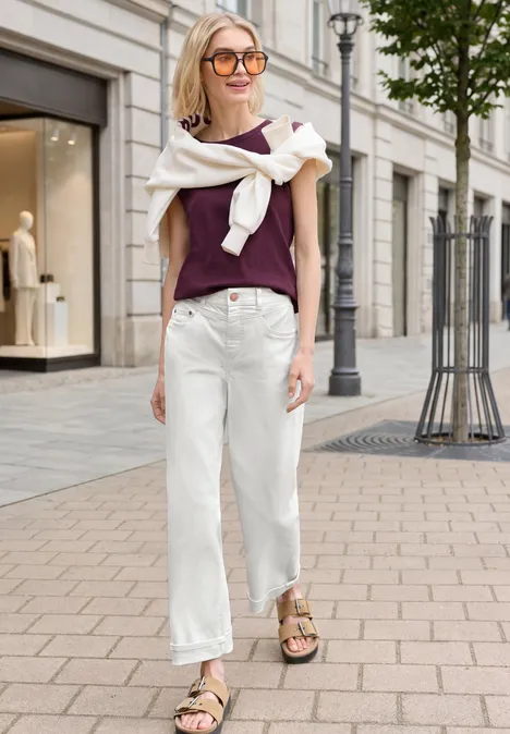 Frau in lila Top, weißer Hose und Sandalen schlendert lässig die Straße entlang.