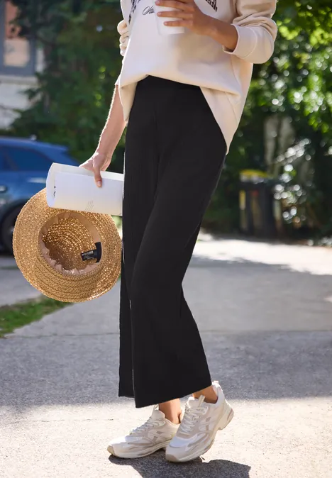 Wide Leg Hose in Rippstruktur Black Eine Person in schwarzen Culottes, weißen Sneakern und mit Strohhut spaziert.