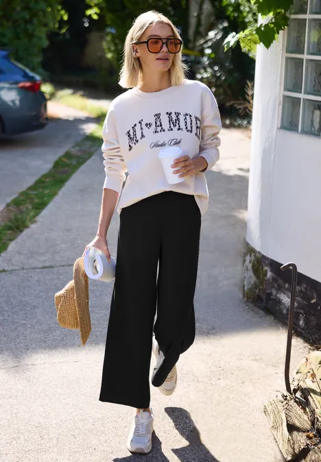 Wide Leg Hose in Rippstruktur Black Frau mit Sonnenbrille, weißem Sweatshirt und Culotte spaziert entspannt einen Gehweg entlang und hält einen Strohhut.