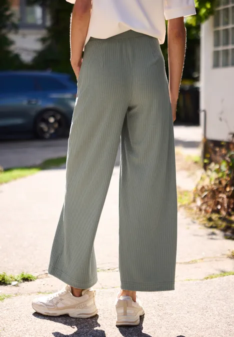 Wide Leg Hose in Rippstruktur sea spray green Rückansicht einer Person in weiten olivgrünen Hosen und weißen Sneakern auf einem sonnigen Bürgersteig.