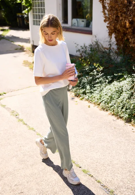 Wide Leg Hose in Rippstruktur sea spray green Junge Frau in weißem T-Shirt und grüner Hose hält Bücher und spaziert auf sonnigem Gehweg.