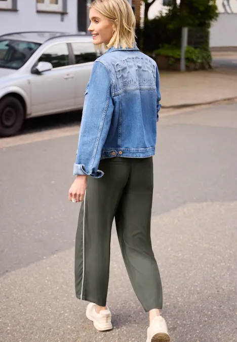Wide Leg Culotte im Loose Fit mit Piping ashen olive Person in blauer Jeansjacke und grünen Jogginghosen mit Seitenstreifen läuft auf der Straße.