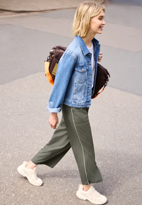 Wide Leg Culotte im Loose Fit mit Piping ashen olive Eine Person in Jeansjacke, weiten grünen Hosen und Sneakern geht auf dem Gehweg und trägt einen braunen Rucksack.