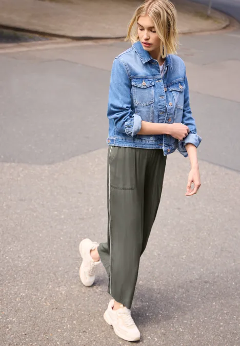 Wide Leg Culotte im Loose Fit mit Piping ashen olive Eine Frau mit Jeansjacke, weiten olivgrünen Hosen und weißen Sneakers steht auf der Straße.