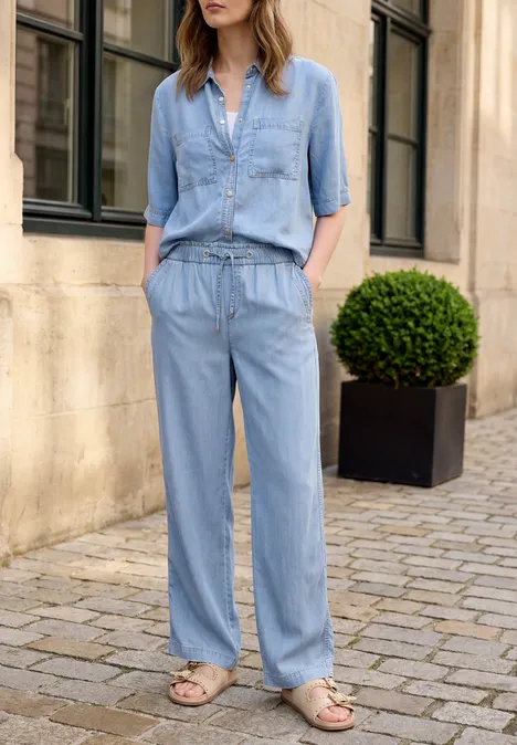 Frau in hellblauem Denim-Jumpsuit mit Taschen und hochgekrempelten Ärmeln, steht in Sandalen auf Pflastersteinen.