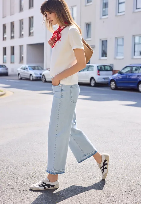 Een jonge vrouw, in spijkerbroek, T-shirt, rood sjaaltje en rugzak, loopt over straat.