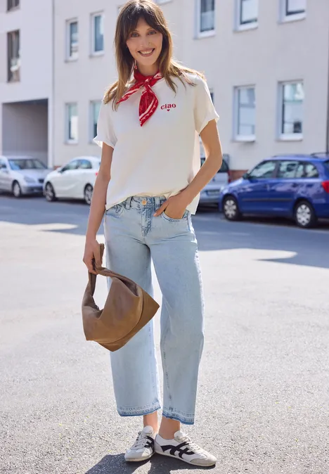 Een vrouw in een wit T-shirt, lichte jeans, slip-on schoenen en rood sjaaltje, met een bruine handtas.