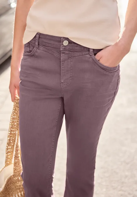 Vrouw in paarse, hoog getailleerde broek met zakken, licht topje en een strooien tas.