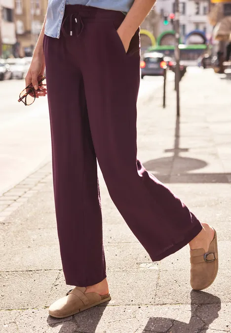 Persoon met wijde paarse broek en beige klompen lopend over een zonnige stadsstraat.