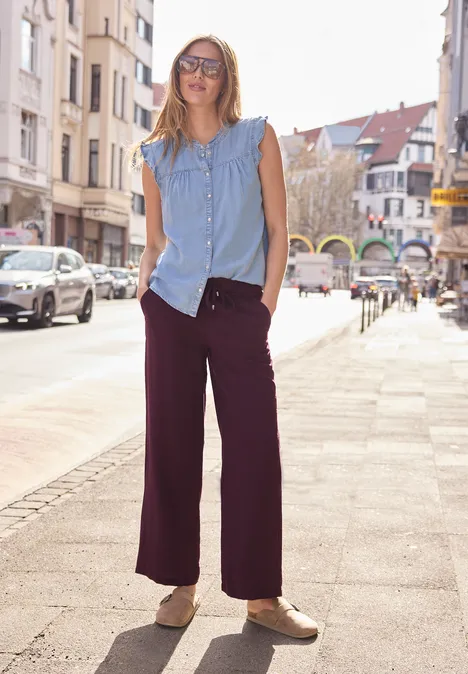 Een persoon staat op een zonnige straat, draagt een lichtblauw mouwloos shirt, wijde paarse broek en lichte schoenen.