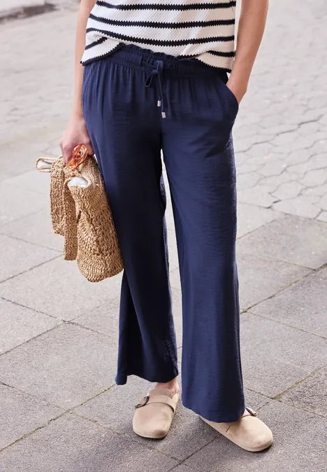 Persoon met wijde marineblauwe broek, gestreept shirt, rieten tas en beige espadrilles op het trottoir.