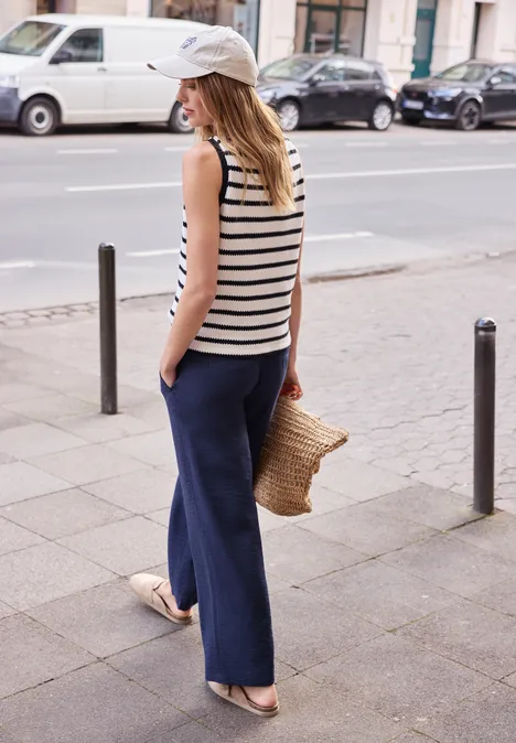 Vrouw in gestreept hemd, brede broek, strooien tas en pet lopend op stoep.