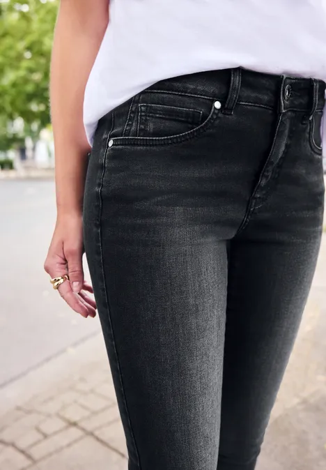 Mid Waist Skinny Leg Jeans im Skinny Fit black washed Person in enger schwarzer Jeans und weißem Oberteil steht draußen auf dem Gehweg.