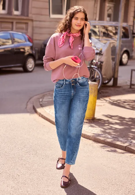 Junge Frau in rosa Oberteil und Jeans mit rosa Handy telefoniert fröhlich beim Spaziergang.