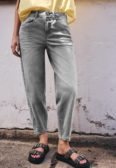Femme en jean ample gris clair et sandales noires à boucles devant un mur écaillé.