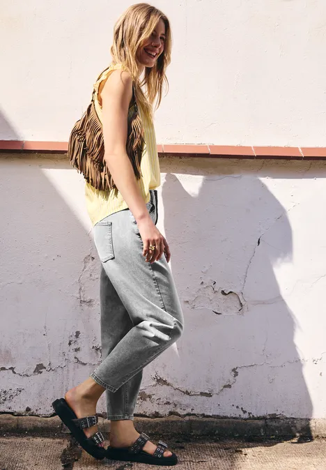 Une jeune femme en top jaune, pantalon gris et sandales noires, appuyée décontractée contre un mur blanc.