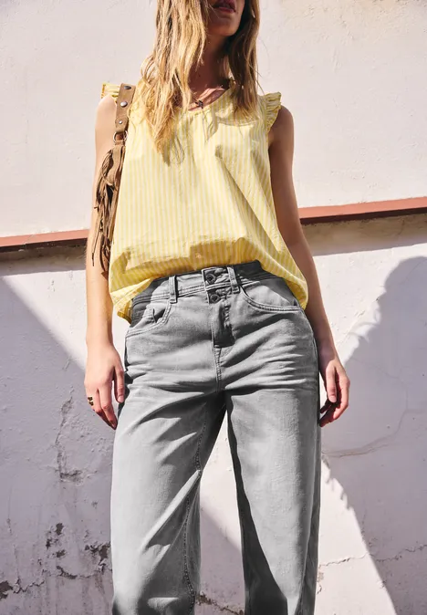 Femme portant un top jaune sans manches et un jean gris devant un mur clair en plein soleil.