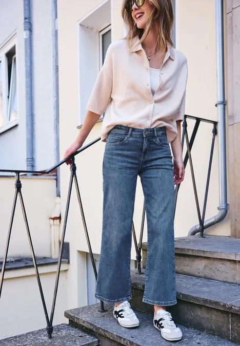 Une femme posant sur un escalier porte une chemise blanche ample, un jean à jambes larges et des baskets blanches.