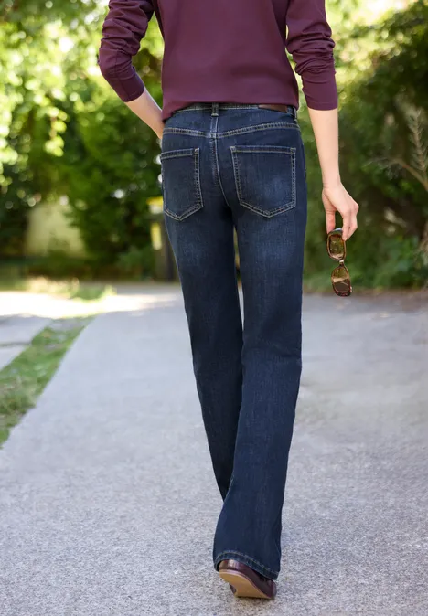 Mid Waist Bootcut Jeans im Slim Fit intense dark blue Person in dunkelblauen Jeans und Pullover geht über einen Gartenweg, hält eine Sonnenbrille