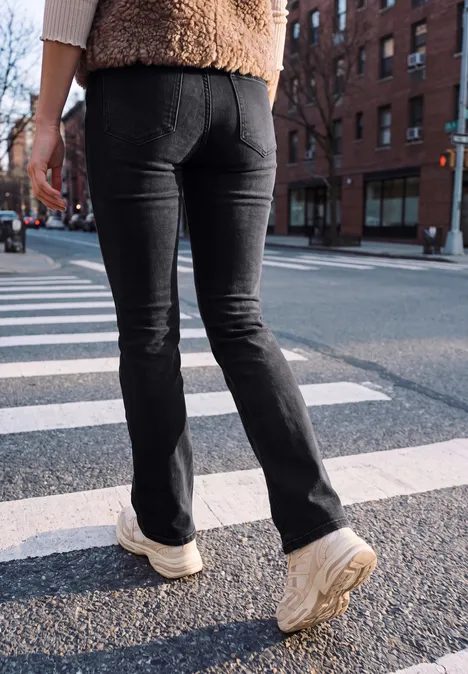 Bootcut-jeans met Mid Waist-taille in Slim Fit authentic black washed Person von hinten überquert Zebrastreifen, trägt schwarze Schlagjeans und helle Sneaker in urbaner Umgebung.
