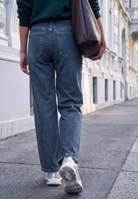 Mid Waist Wide Leg Jeans im Casual Fit dark blue soft washed Person in blauen Jeans, Turnschuhen und Tasche läuft auf Gehweg entlang eleganter Häuser.