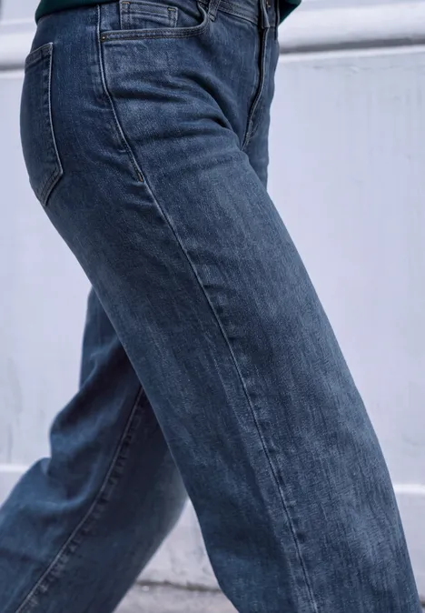 Mid Waist Wide Leg Jeans im Casual Fit dark blue soft washed Seitliche Ansicht einer Person in dunkelblauen Jeanshose vor weißer Wand.