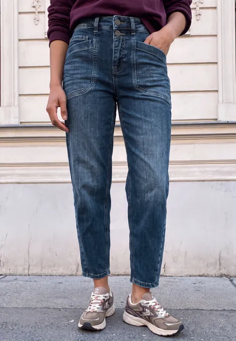 Person trägt knöchellange blaue Jeans und beige Sneaker vor heller Holzfassade.