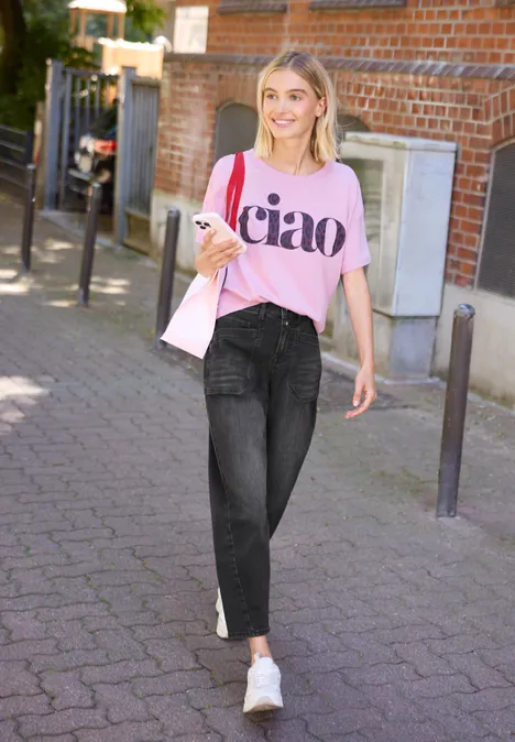 Frau in pinkem "ciao"-Shirt und Jeans trägt Tasche und geht spazieren.