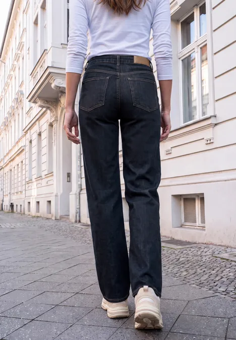 Mid Waist Wide Leg Jeans im Loose Fit rinsed blue washed Eine Person in schwarzen Jeans und weißen Sneakers geht auf einem Gehweg vor Häuserfassaden spazieren.
