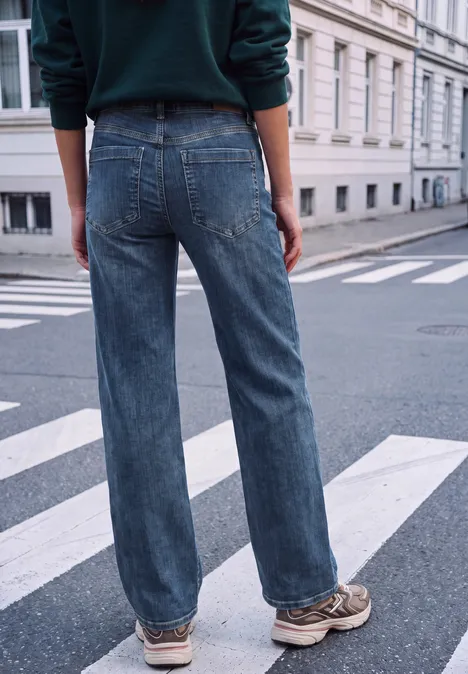 Mid Waist Wide Leg Jeans im Loose Fit mid blue heavy washed Person steht auf Zebrastreifen, trägt weite blaue Jeans und Sneaker vor heller Hausfassade.