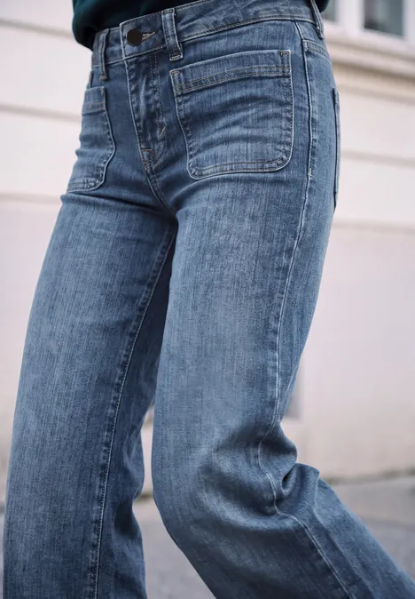Mid Waist Wide Leg Jeans im Loose Fit mid blue heavy washed Seitliche Nahaufnahme einer Person in lockeren, blauen Jeans mit aufgesetzten Taschen vor hellem Hintergrund.