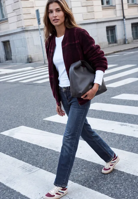Mid Waist Straight Leg Jeans im Casual Fit dark blue random washed Eine junge Frau in Jeans, weißem Shirt und bordeauxrotem Cardigan überquert eine städtische Straße mit Handtasche.