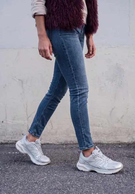 Mid Waist Slim Leg Jeans im Casual Fit medium blue washed Eine Person in Jeans und grauen Sneakers geht vor einer weißen Wand entlang.