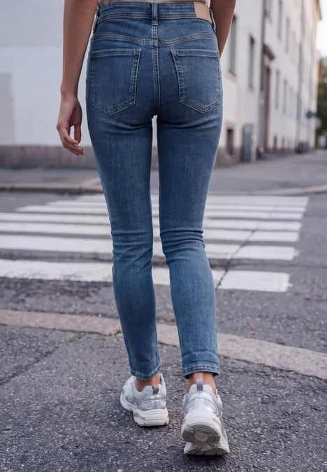 Mid Waist Slim Leg Jeans im Casual Fit medium blue washed Person in engen blauen Jeans und Sneakers, von hinten auf einem Zebrastreifen in der Stadt.