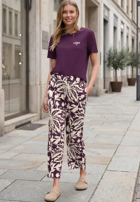 Een vrouw in een paars t-shirt en bloemige broek staat ontspannen op een stedelijke straat.