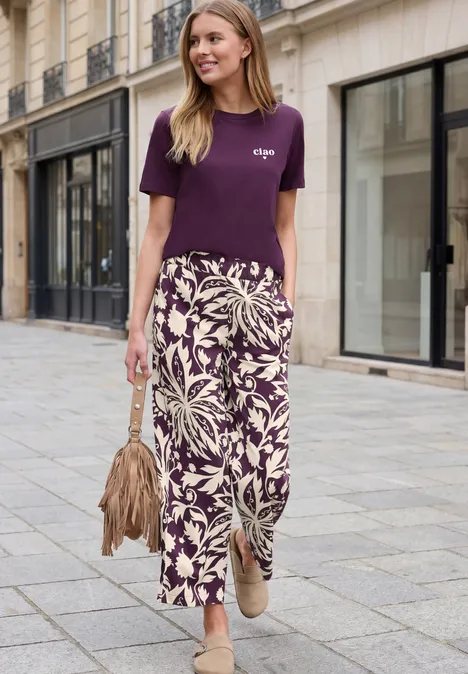Vrouw in paarse T-shirt en licht gekleurde maxirok met bladprint wandelt door de straat.