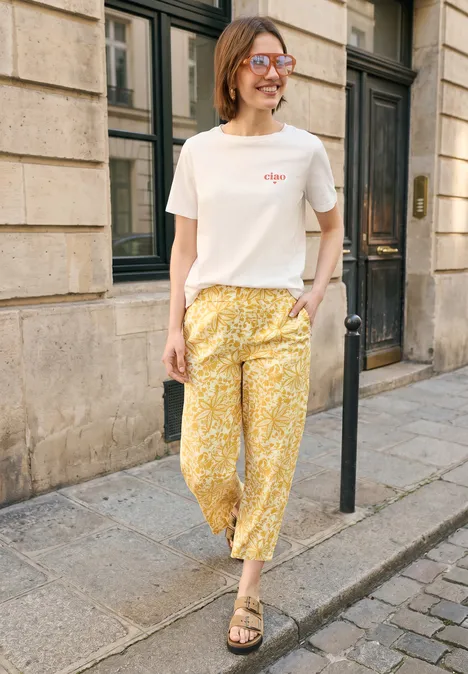 Persoon in wit T-shirt en gele bloemenbroek staat op de stoep voor een stenen gevel.