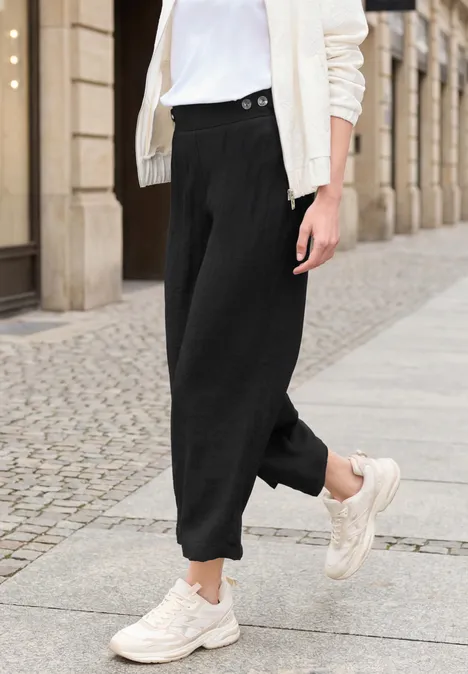 Person trägt schwarze, weite Hose und weiße Sneaker auf gepflastertem Gehweg